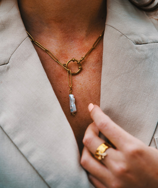 Collier fermoir marin en acier inoxydable doré avec une longue perle d'eau douce. Ce collier est en maille ovale et est porté sur un décolleté avec une veste tailleur.